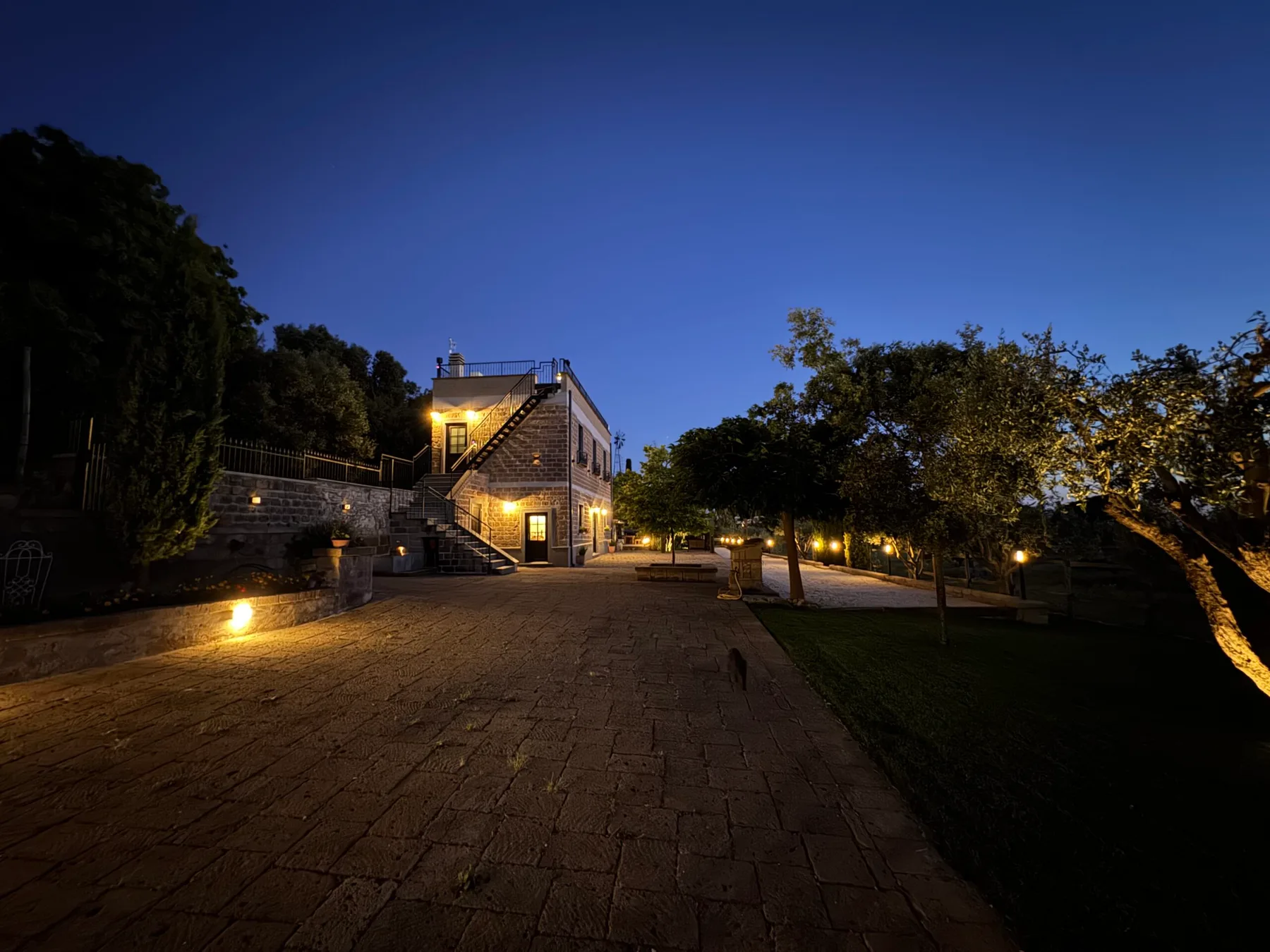 Espacio representativo del hotel: Un patio empedrado al anochecer, con un edificio iluminado y árboles.
