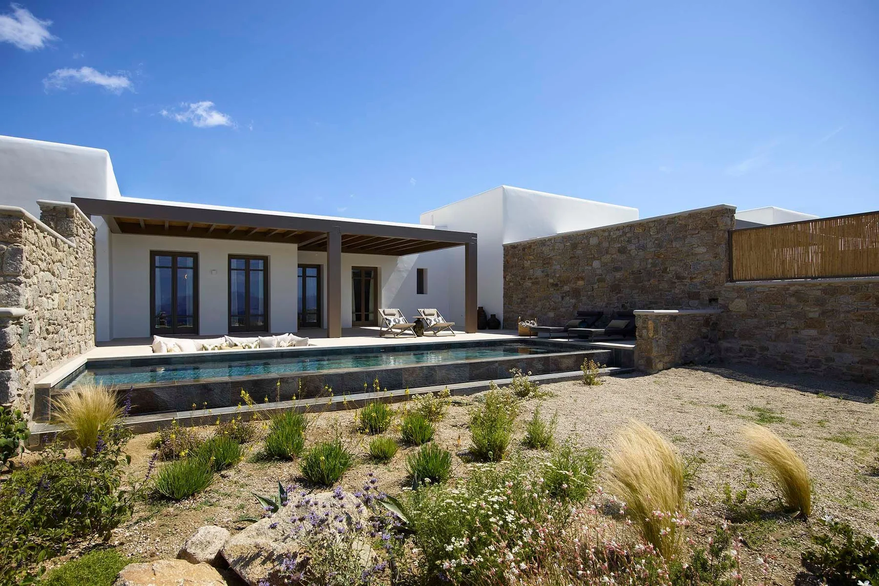 Un patio moderno da a una larga piscina exterior junto a muros de piedra.