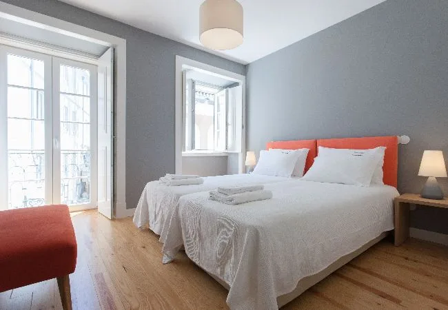Un dormitorio con una cama blanca, un cabecero naranja y ventanas altas.