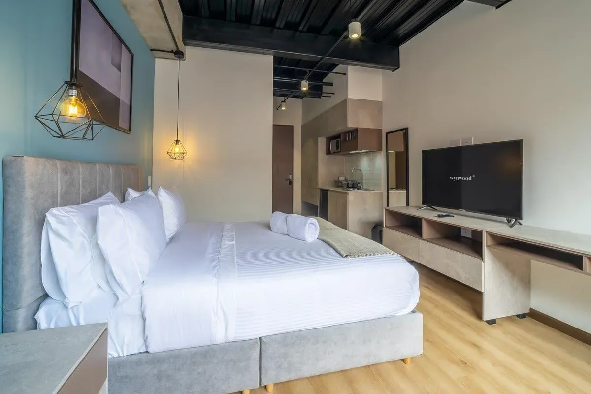 A modern bedroom has a white bed, a wall-mounted television, and a compact kitchenette.