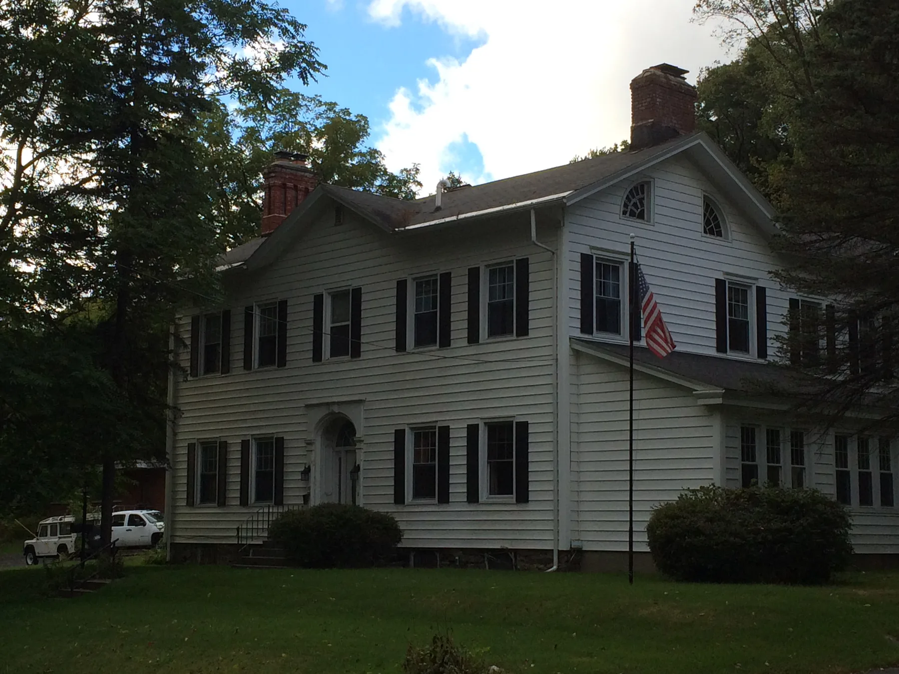 Representative hotel space: A large white house with dark shutters stands among trees beside a lawn.