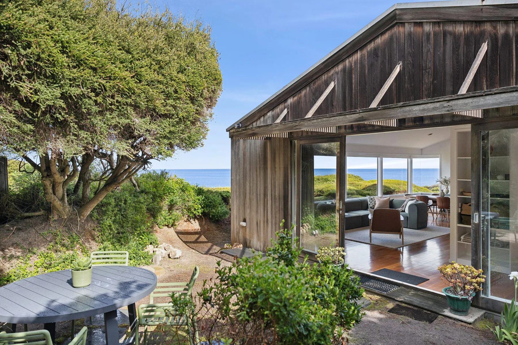 A modern timber house opens onto a garden patio, with the sea visible beyond.