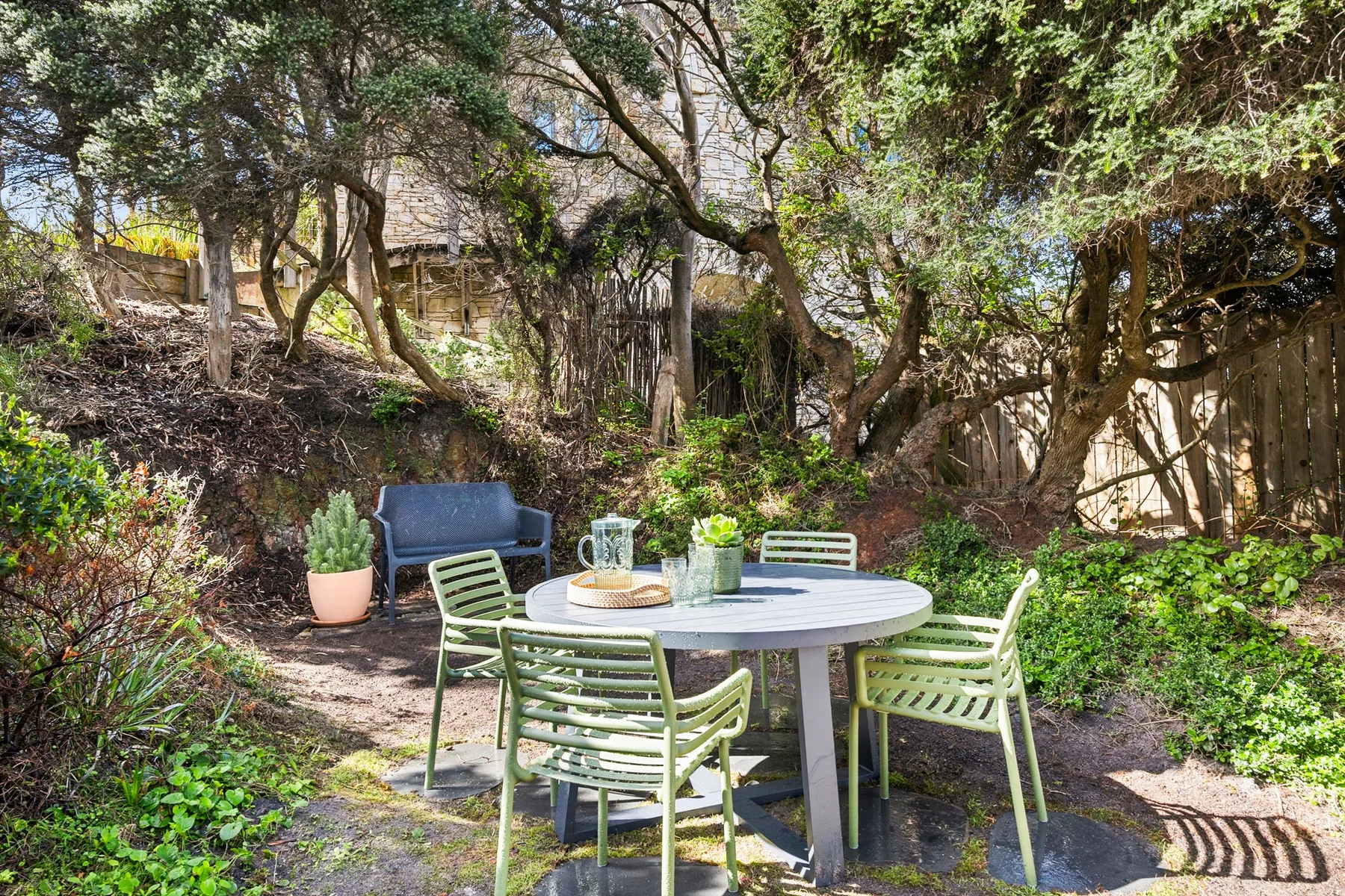 A garden patio with a round table and green seating beneath leafy trees.