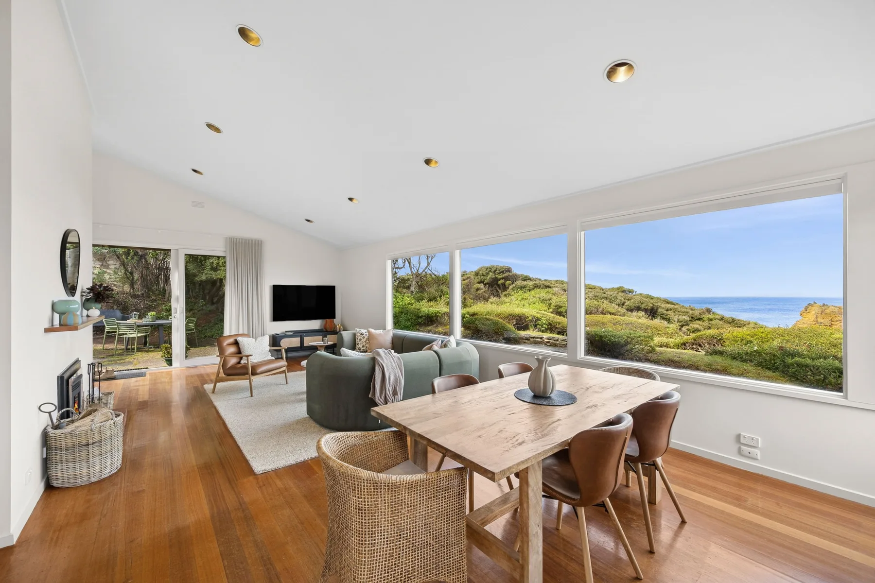 A bright living and dining room with large windows overlooking greenery and the ocean.