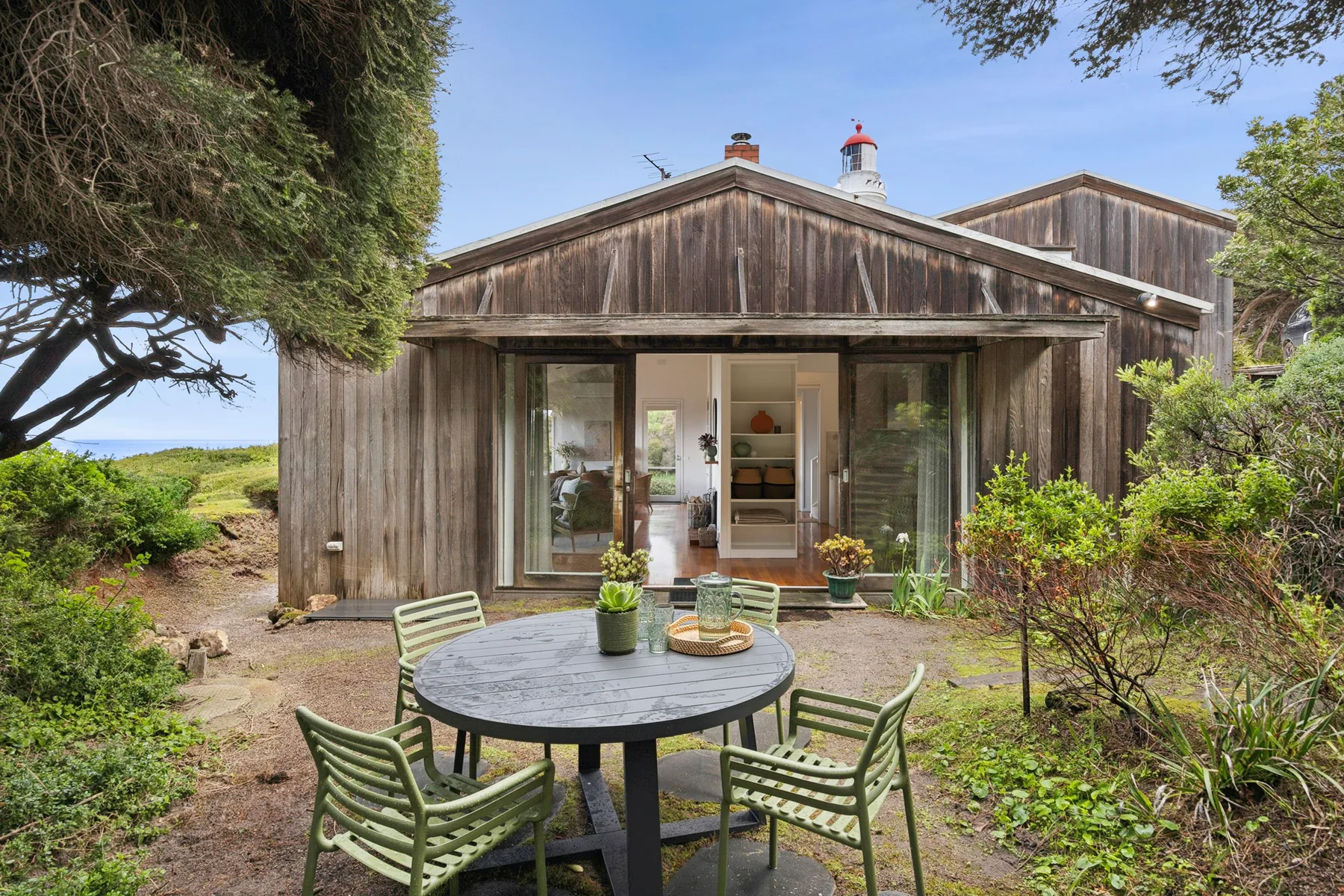 A wooden house with glass doors opens onto a garden patio with outdoor seating.