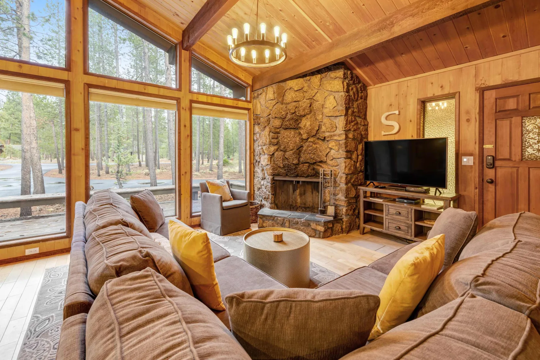 A living room with seating, a stone fireplace, and large windows overlooking trees.