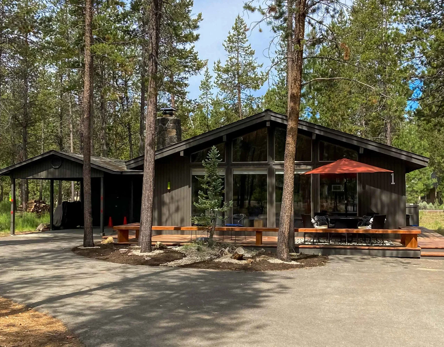 A modern cabin sits among tall pine trees, with a red patio umbrella outside.