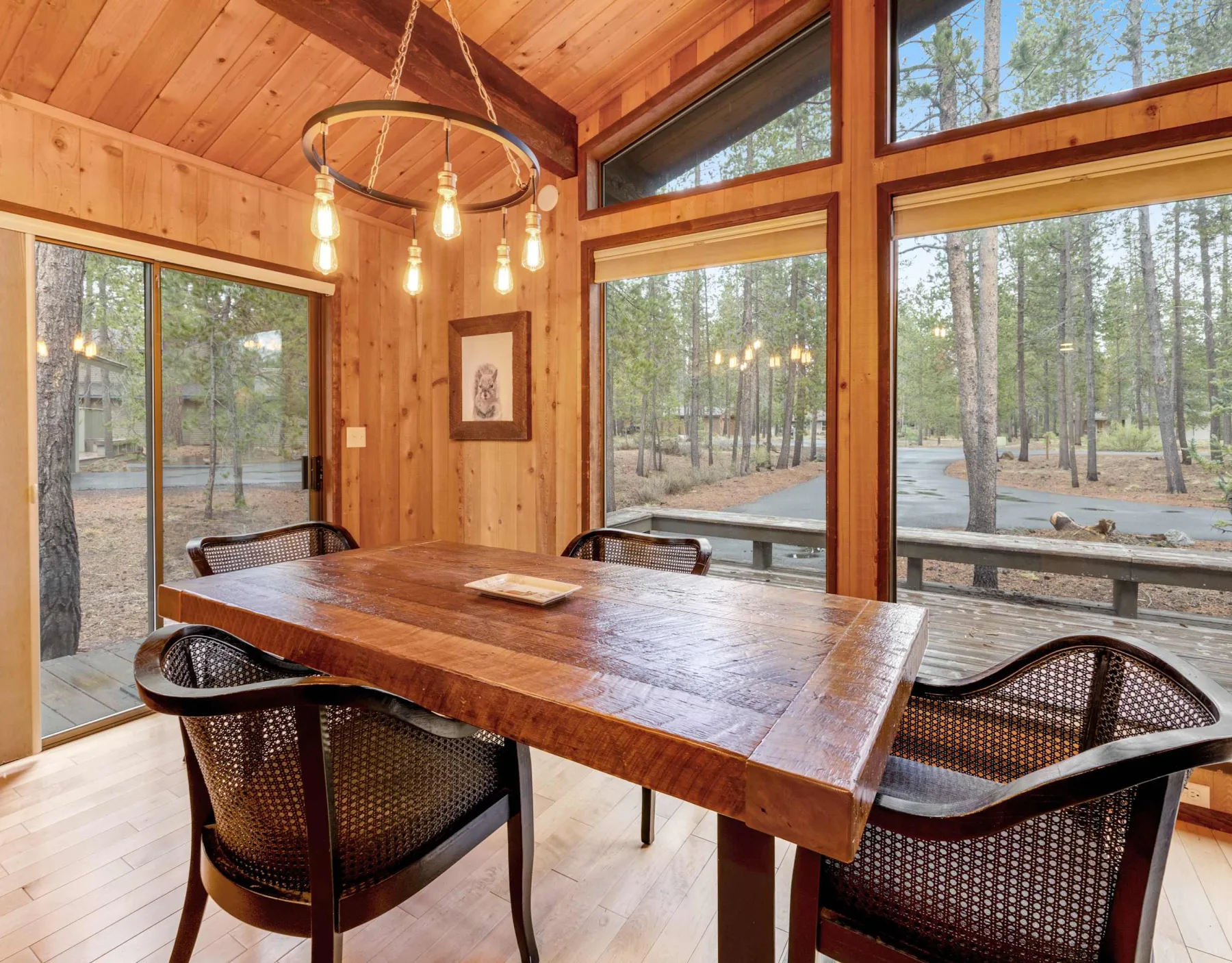 A wooden dining table and seating sit beside large windows overlooking trees.