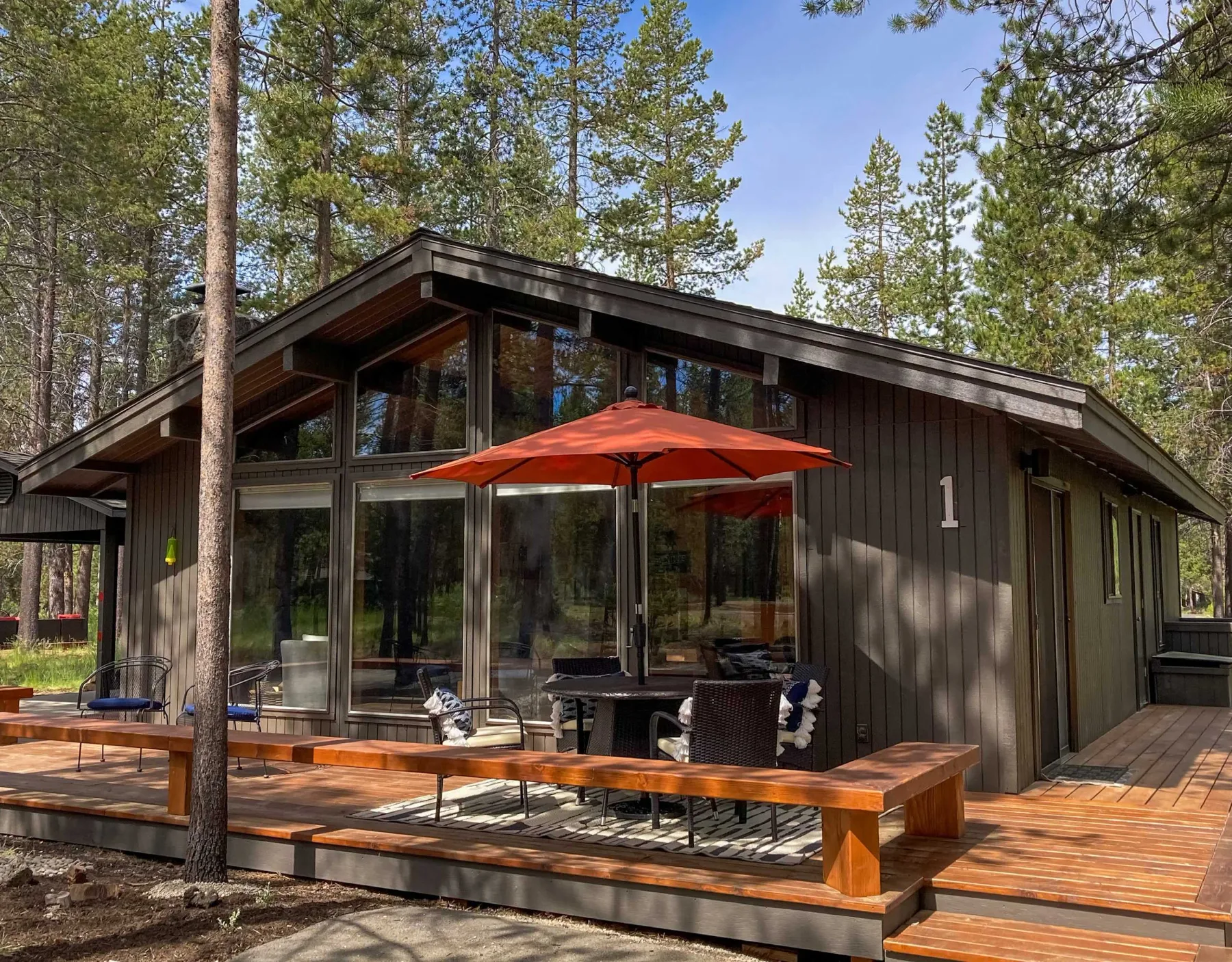 A modern cabin with a deck, outdoor seating, and a red umbrella among pine trees.