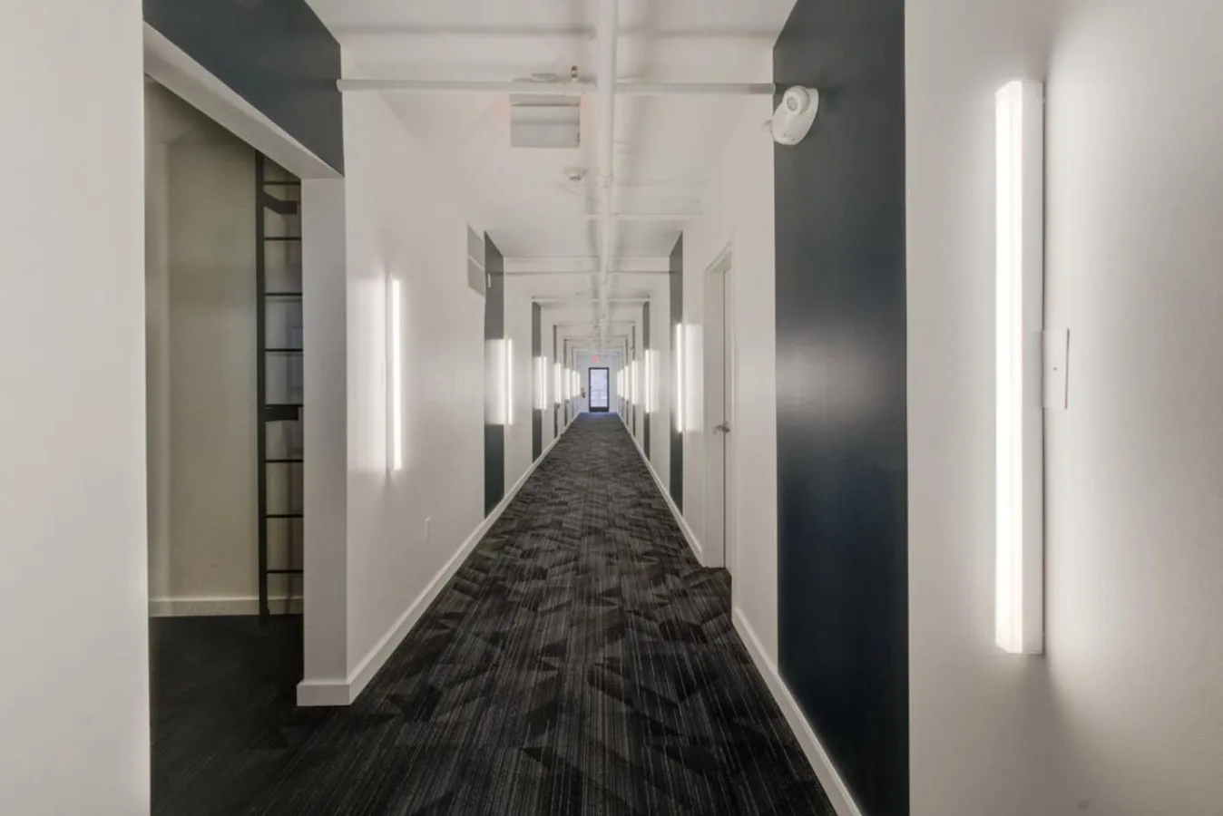 Zona común: Un largo pasillo interior con alfombra oscura estampada y luces en las paredes.