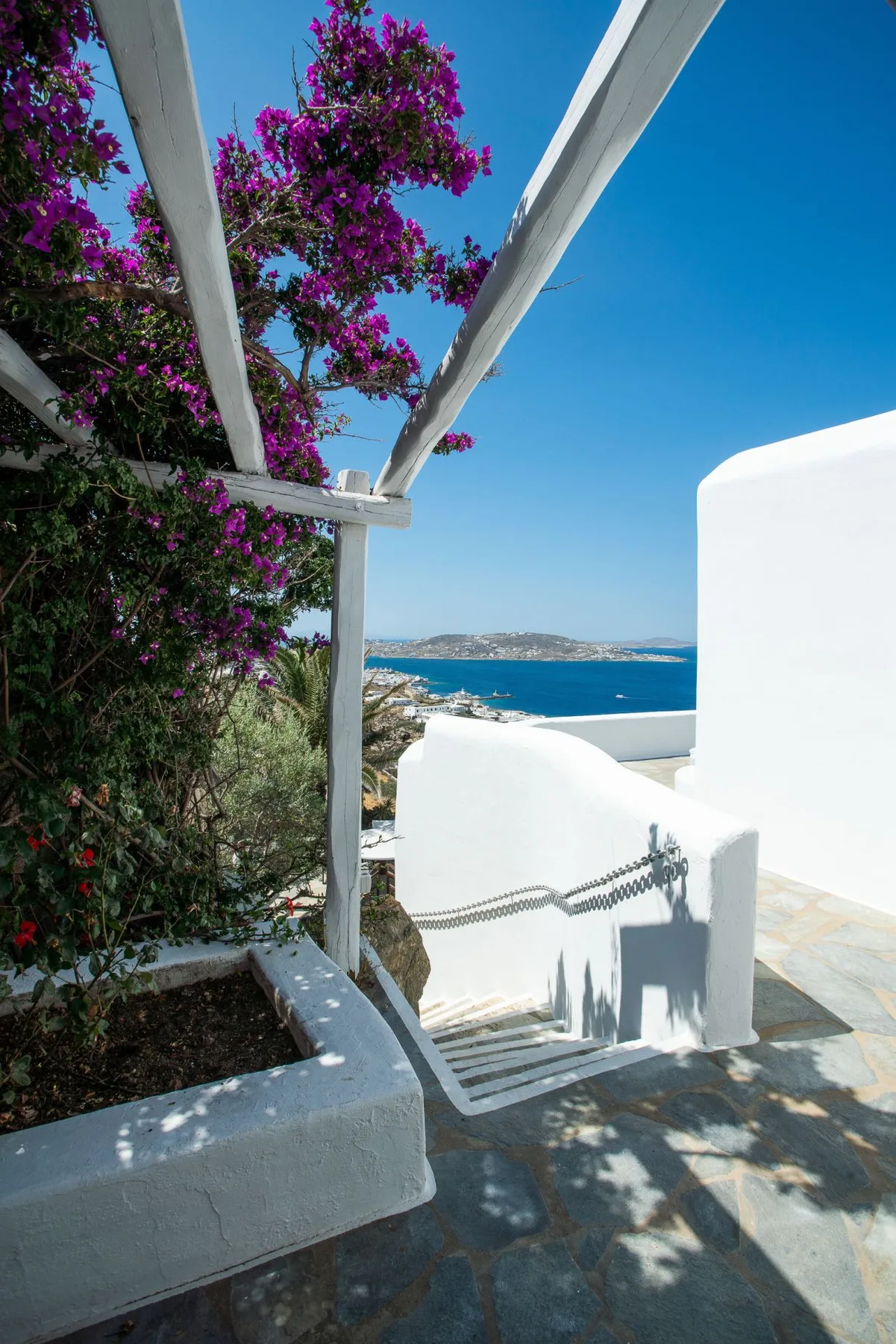 A white outdoor stairway descends beside flowering vines toward a view of the sea.