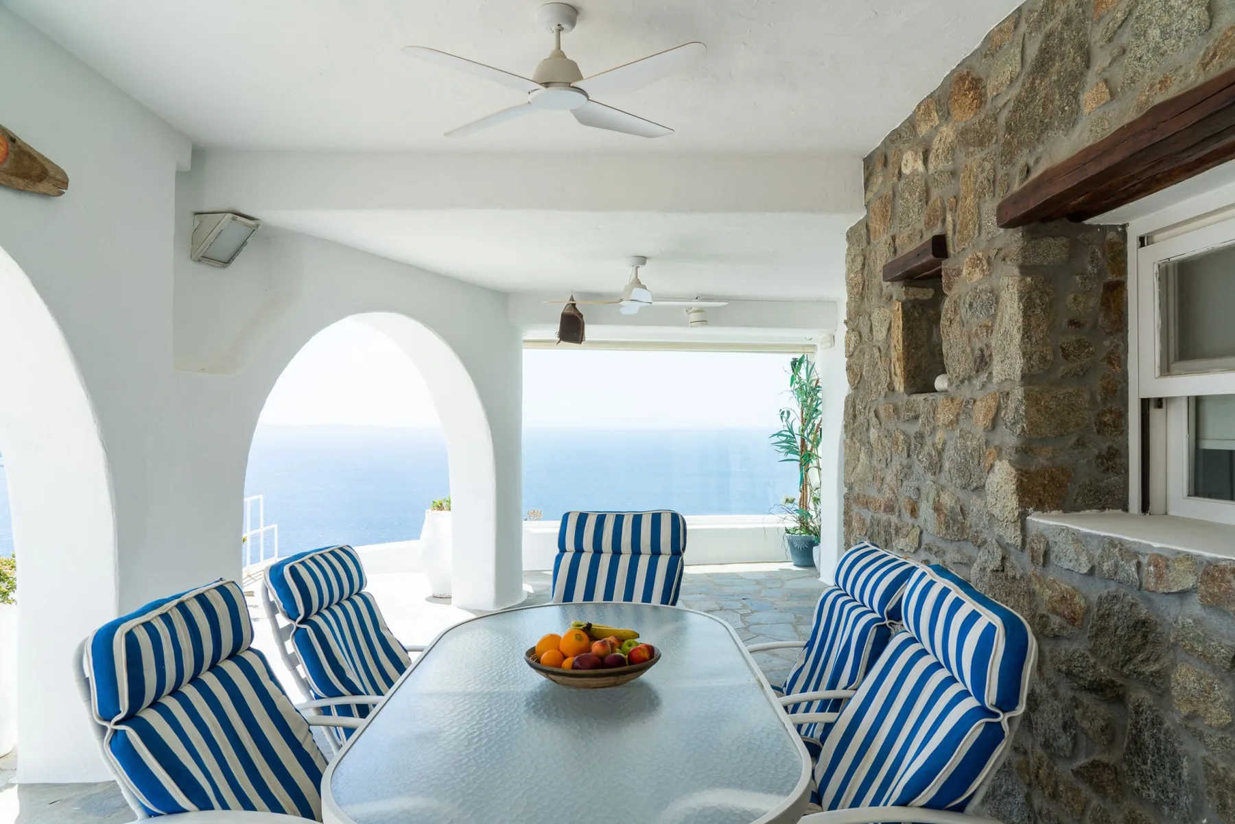A covered patio with striped seating and a table overlooks the sea.