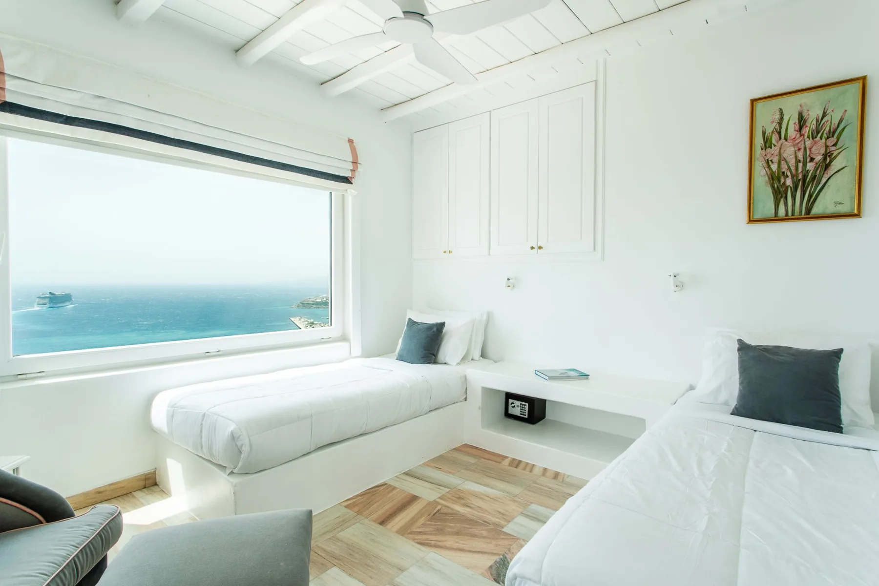 A bright bedroom with two beds beside a large window overlooking the ocean.
