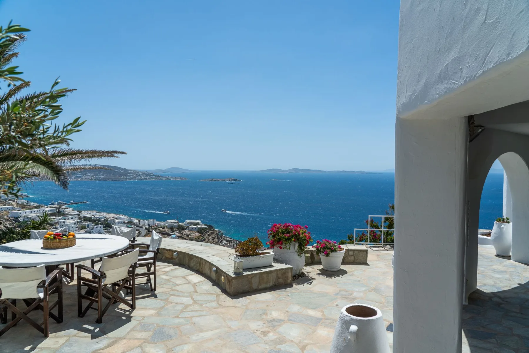 A seaside terrace overlooks the blue water, with outdoor seating and flowering plants.