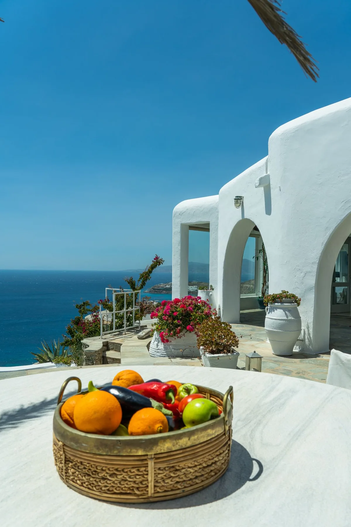A basket of fruit sits on a table beside a white seaside terrace overlooking the water.