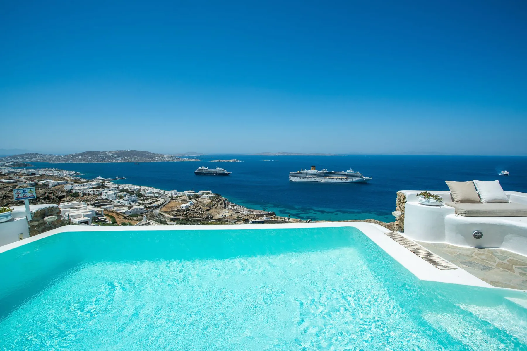 A turquoise pool overlooks the sea and cruise ships under a clear sky.