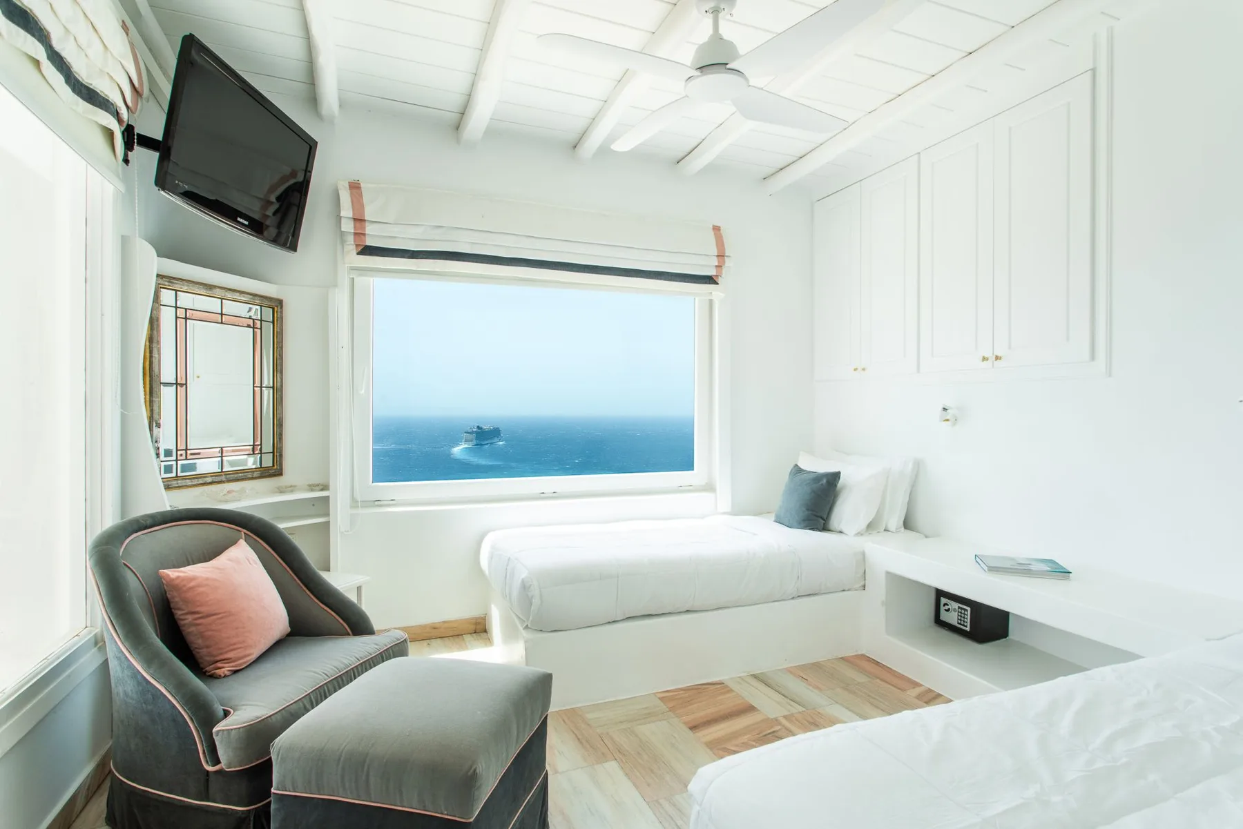 A bright bedroom with a bed beside a large window overlooking the sea.