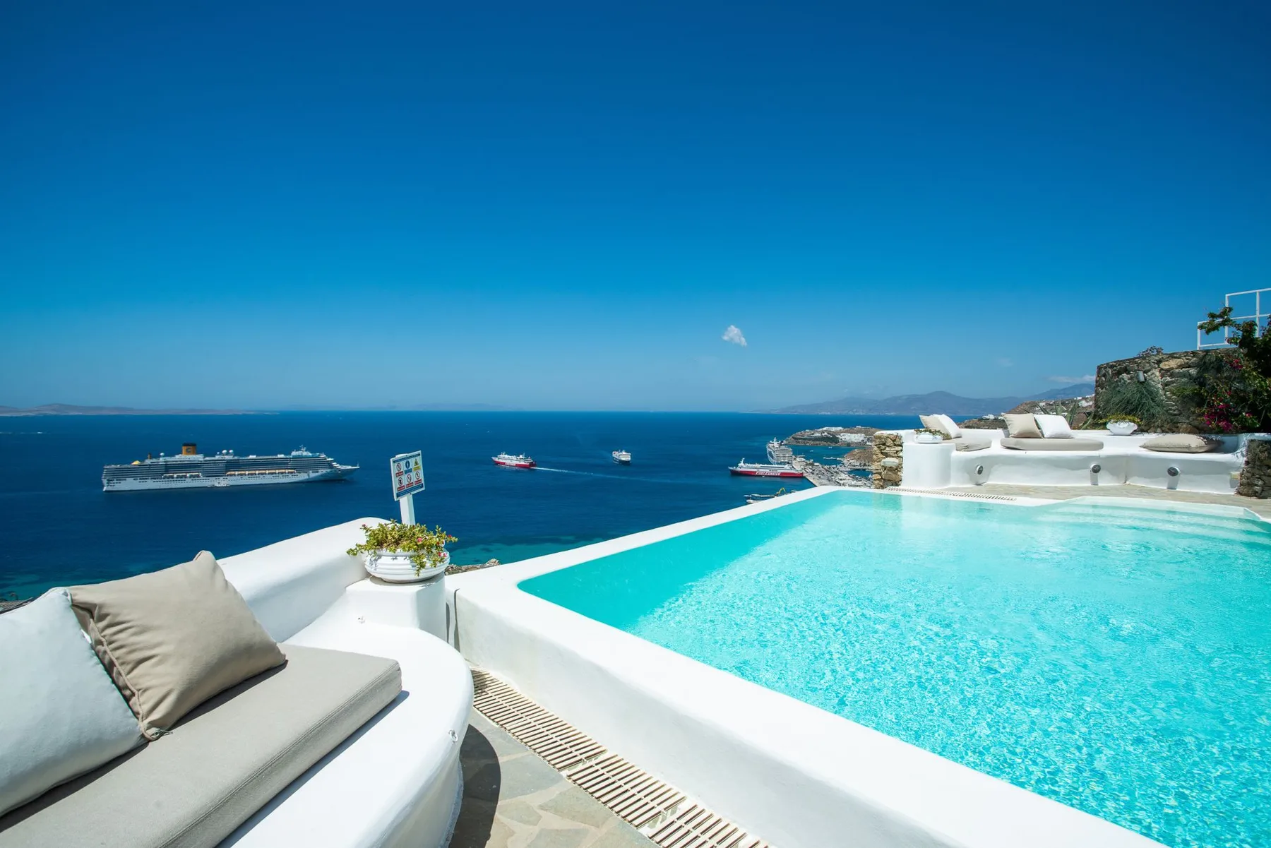 A turquoise swimming pool overlooks the sea, with cushioned seating beside it.