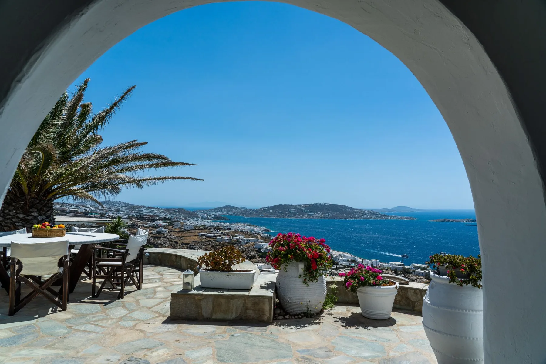A stone terrace overlooks the sea through a large arched opening, with potted flowers nearby.