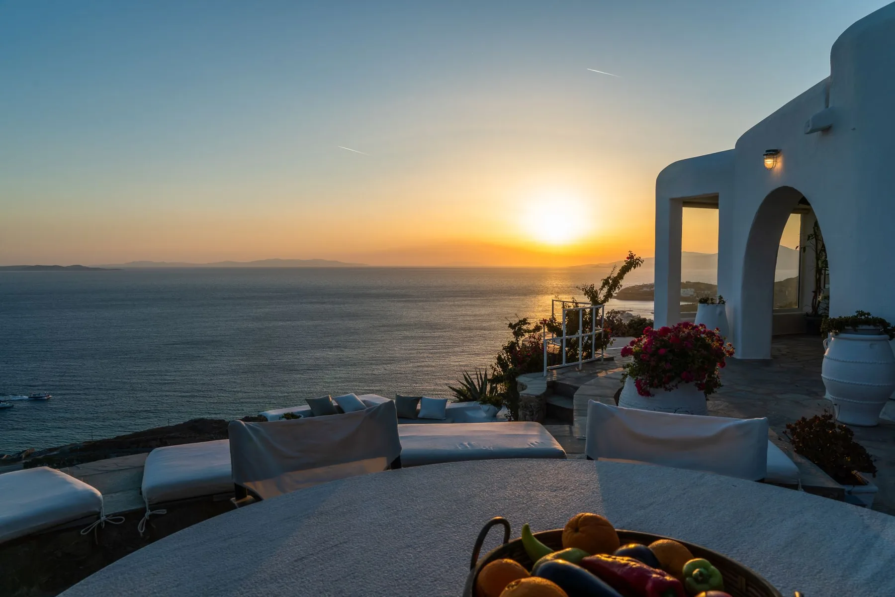 A seaside terrace at sunset overlooks the water, with outdoor seating in the foreground.