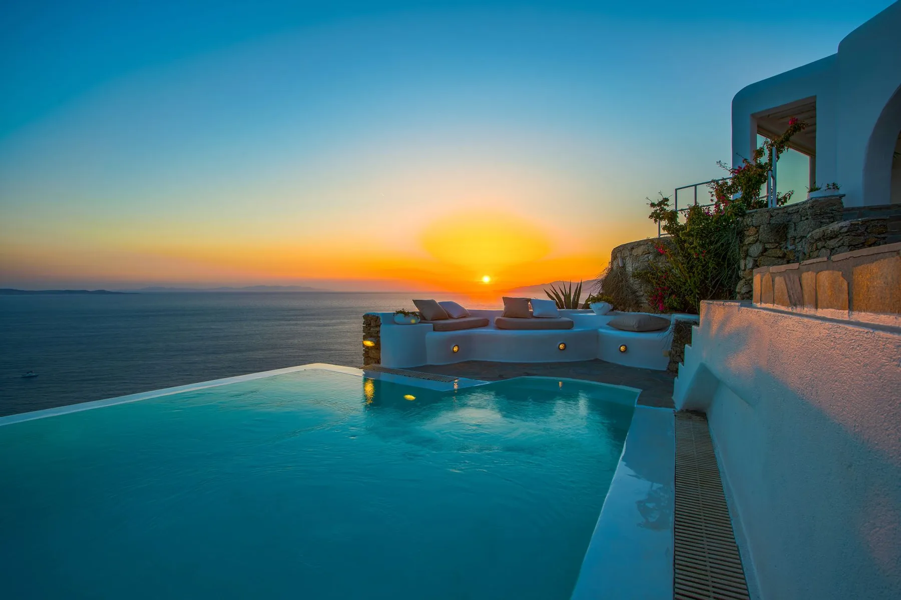 An infinity pool overlooks the sea at sunset beside a building.