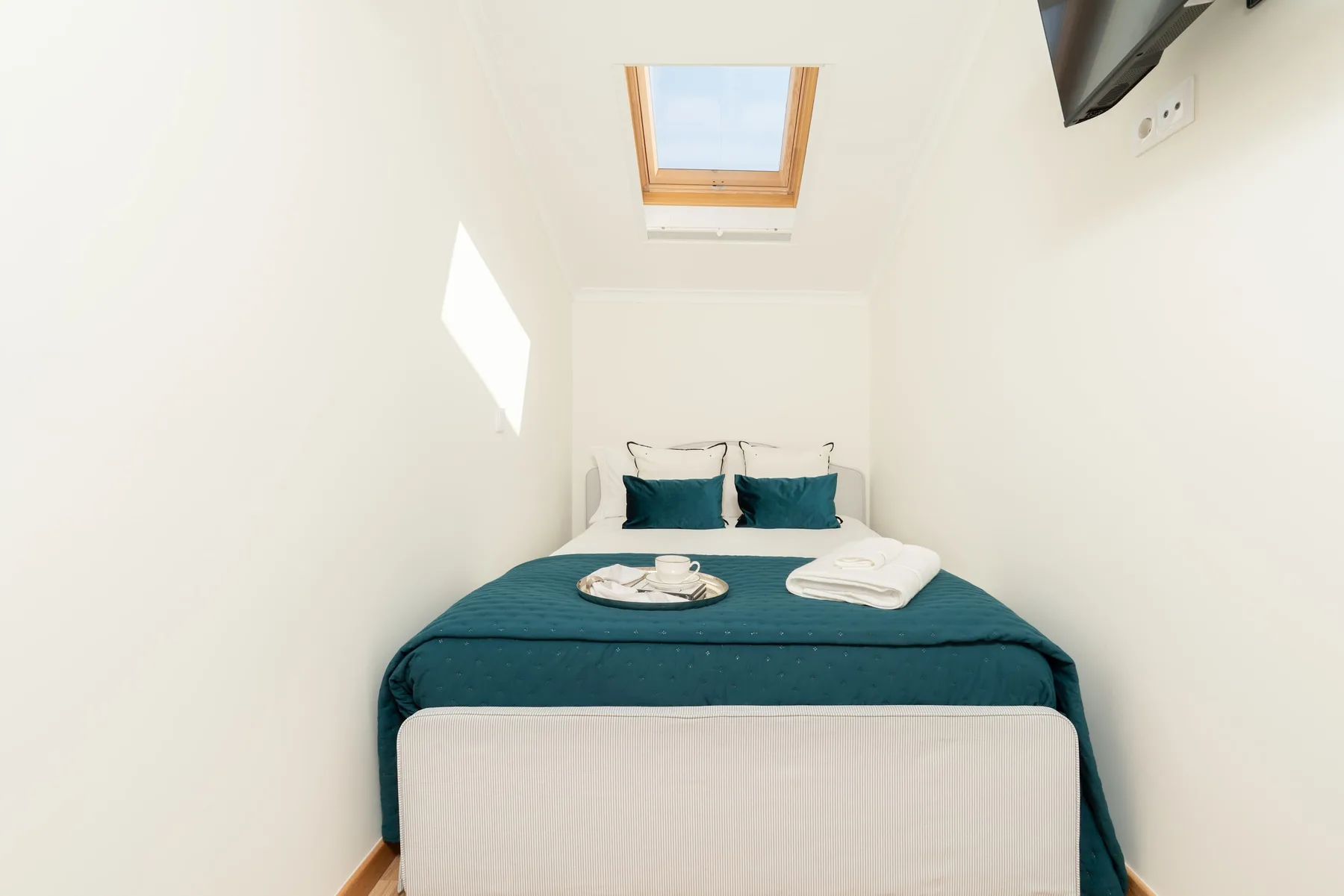 Un dormitorio pequeño con una cama vestida de color verde azulado bajo una claraboya.