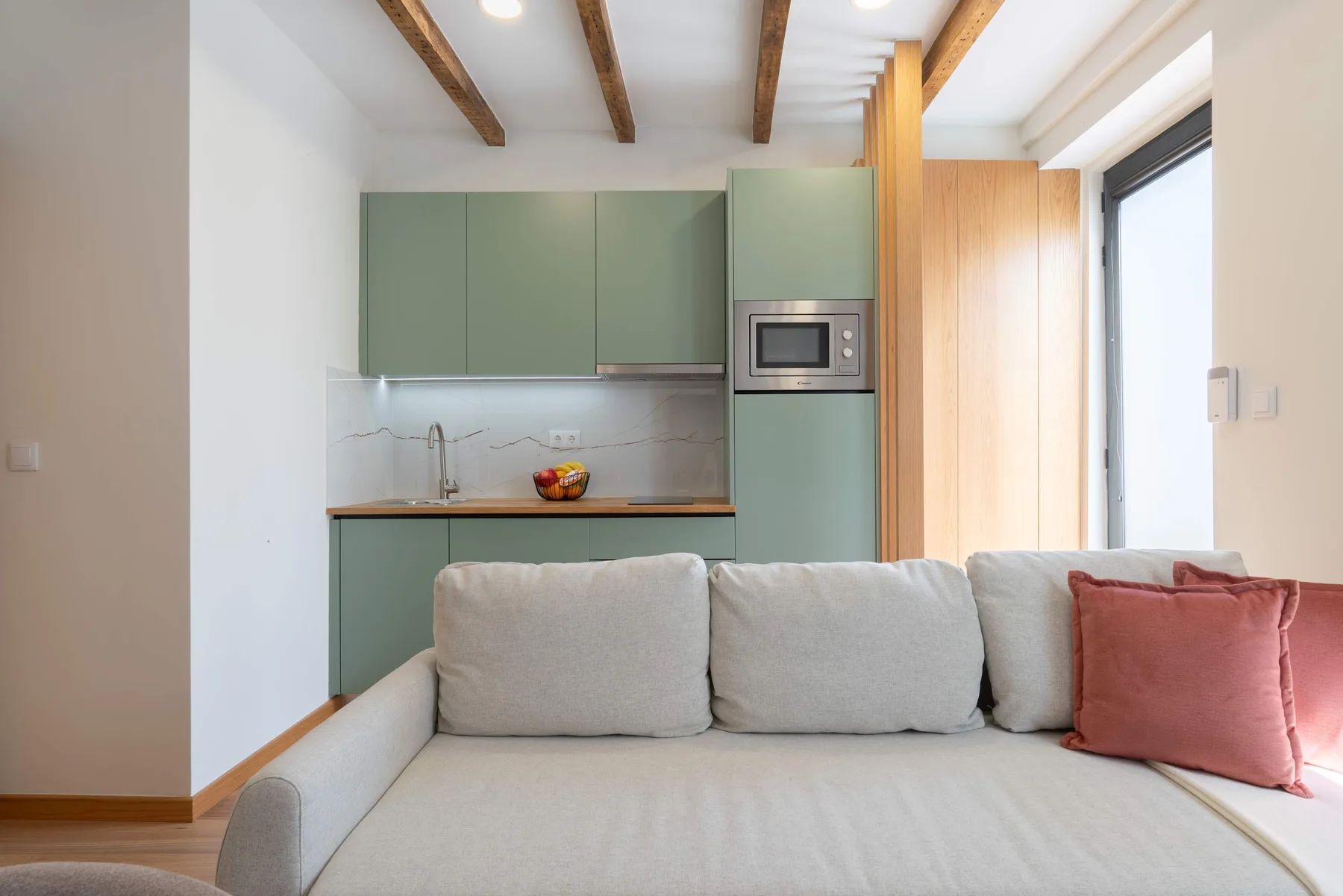 Una luminosa sala de estar con un sofá claro frente a una pequeña cocina.