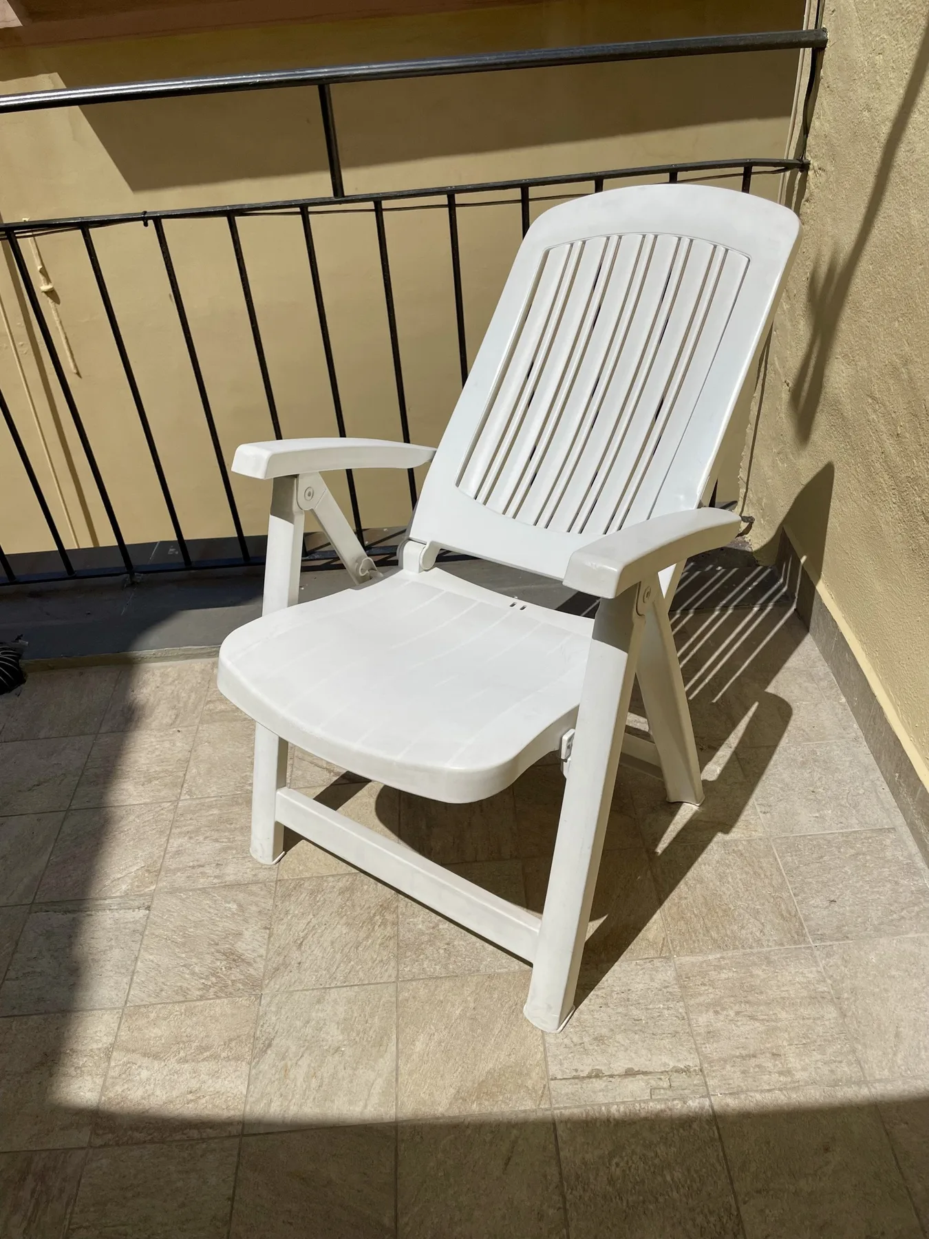 A white plastic chair sits on a sunlit tiled balcony beside a black railing.