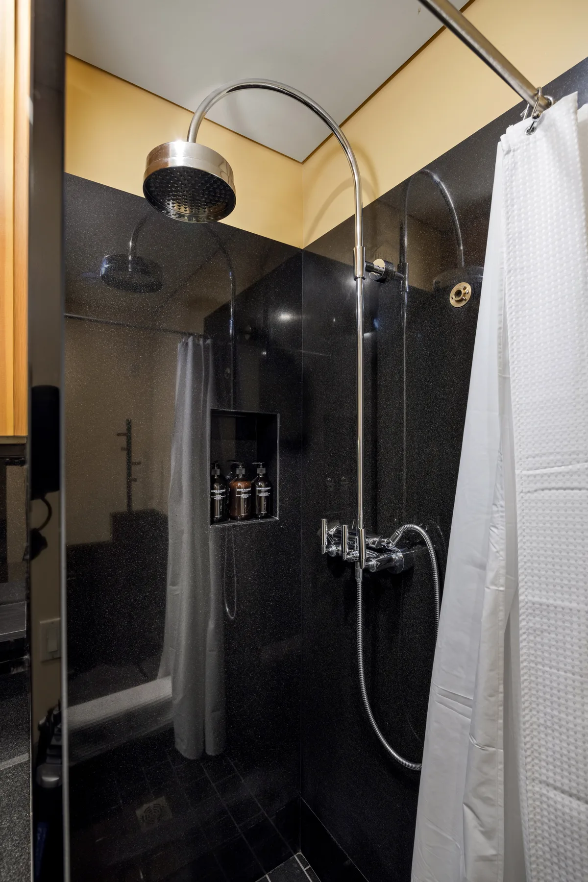 A dark tiled shower area has a curved showerhead and a white curtain.