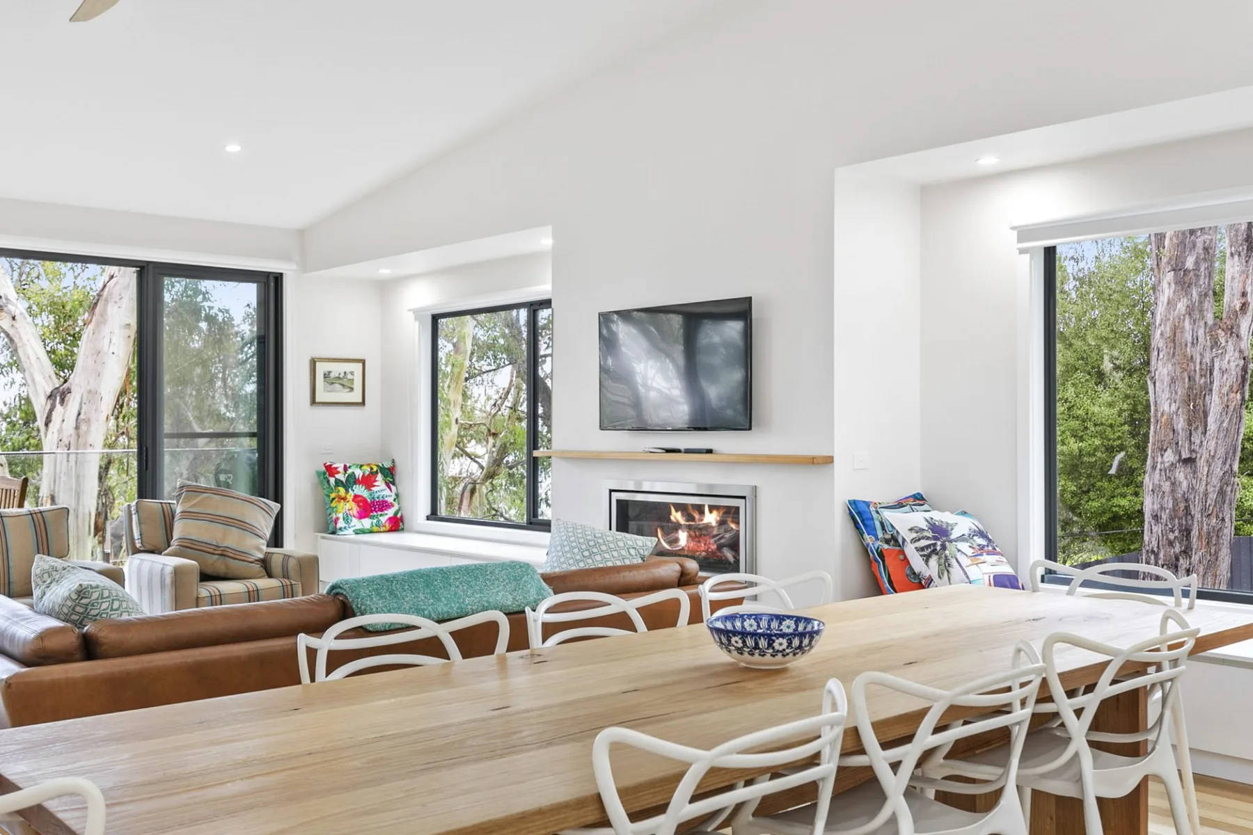 Una luminosa sala de estar y comedor con chimenea, televisor y grandes ventanas.