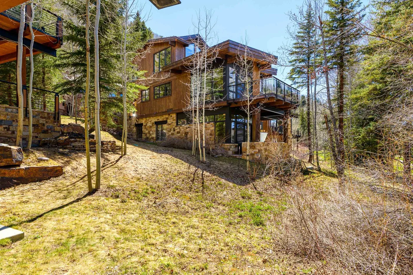 A modern wood-and-stone house stands among trees on a sloping clearing.