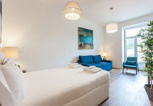 Un dormitorio luminoso con una cama grande, asientos azules y una silla junto a la ventana.