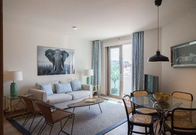 A bright living and dining area has seating, a glass table, and tall windows.