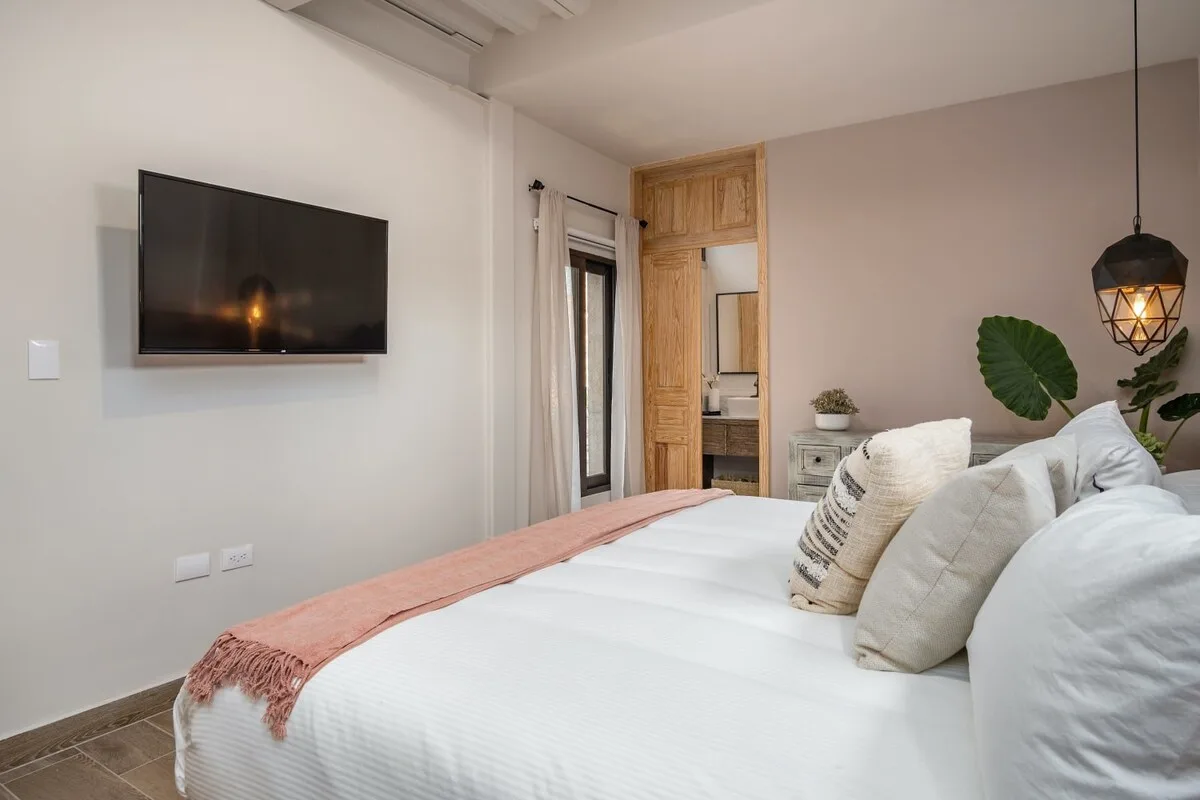 A bright bedroom with a neatly made bed, wall-mounted television, and wooden doorway.