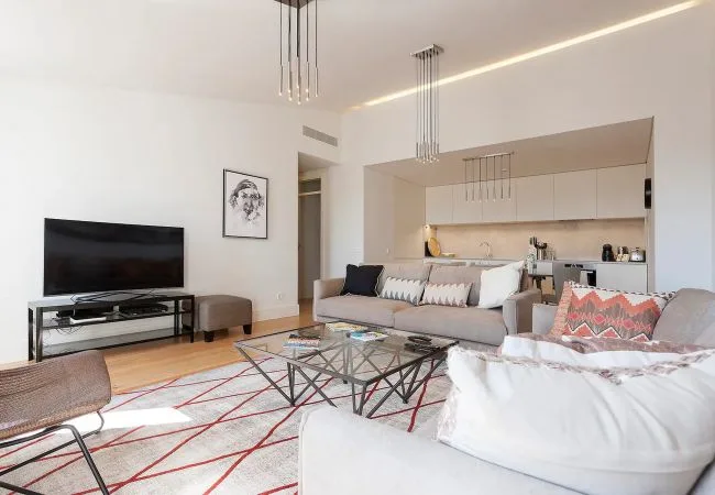 A bright living room with seating, a glass coffee table, and an open kitchen.