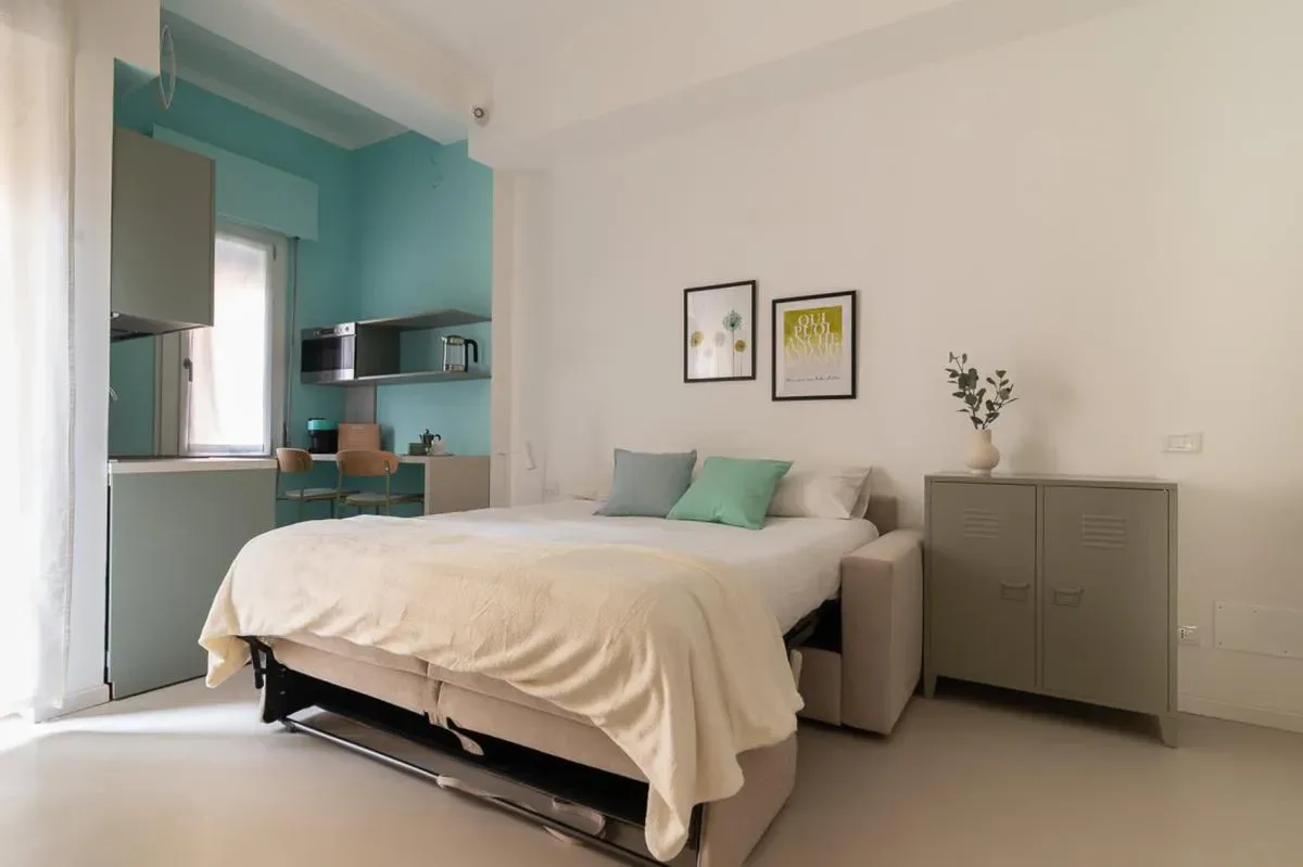 Un dormitorio con una cama, asientos junto a ella y una cocina pequeña turquesa al fondo.