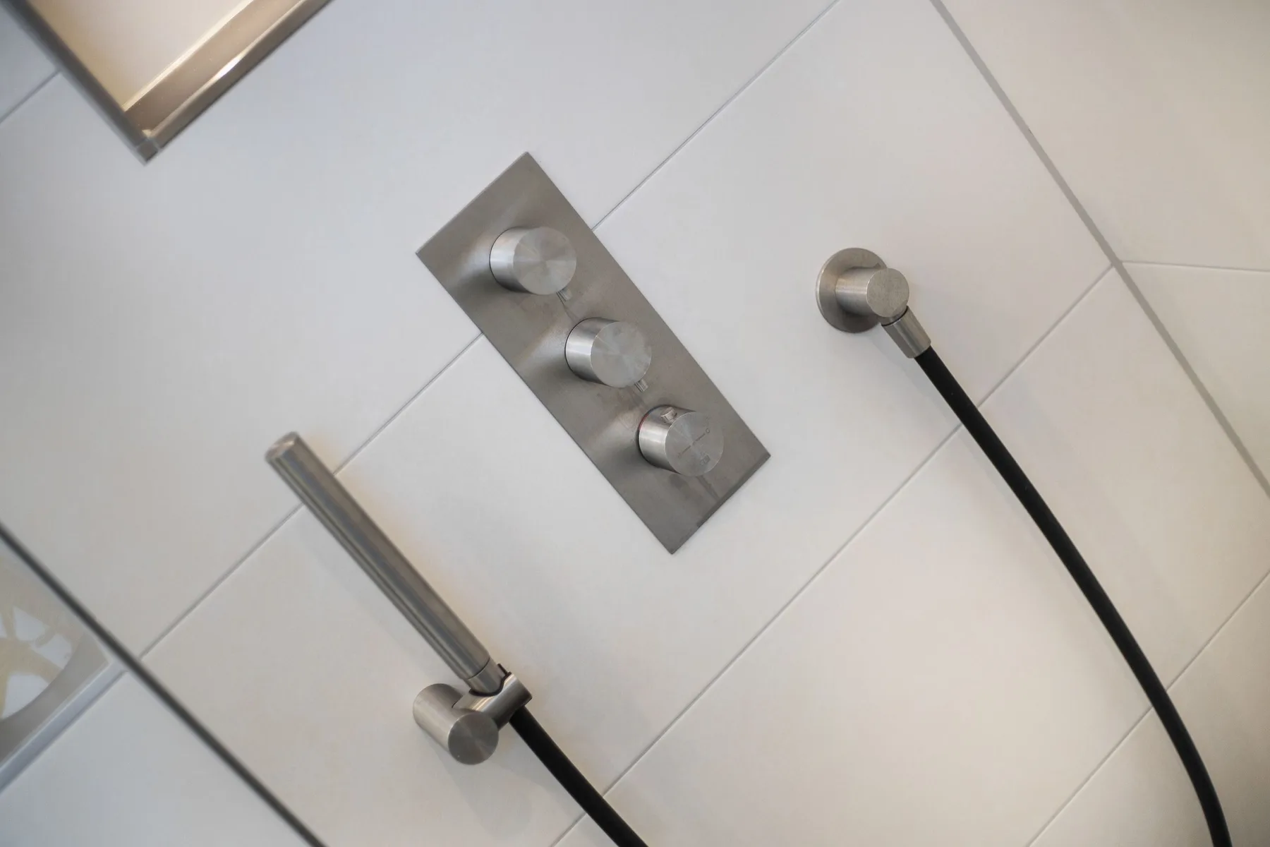 A tiled shower wall with metal controls and handheld shower fixtures.