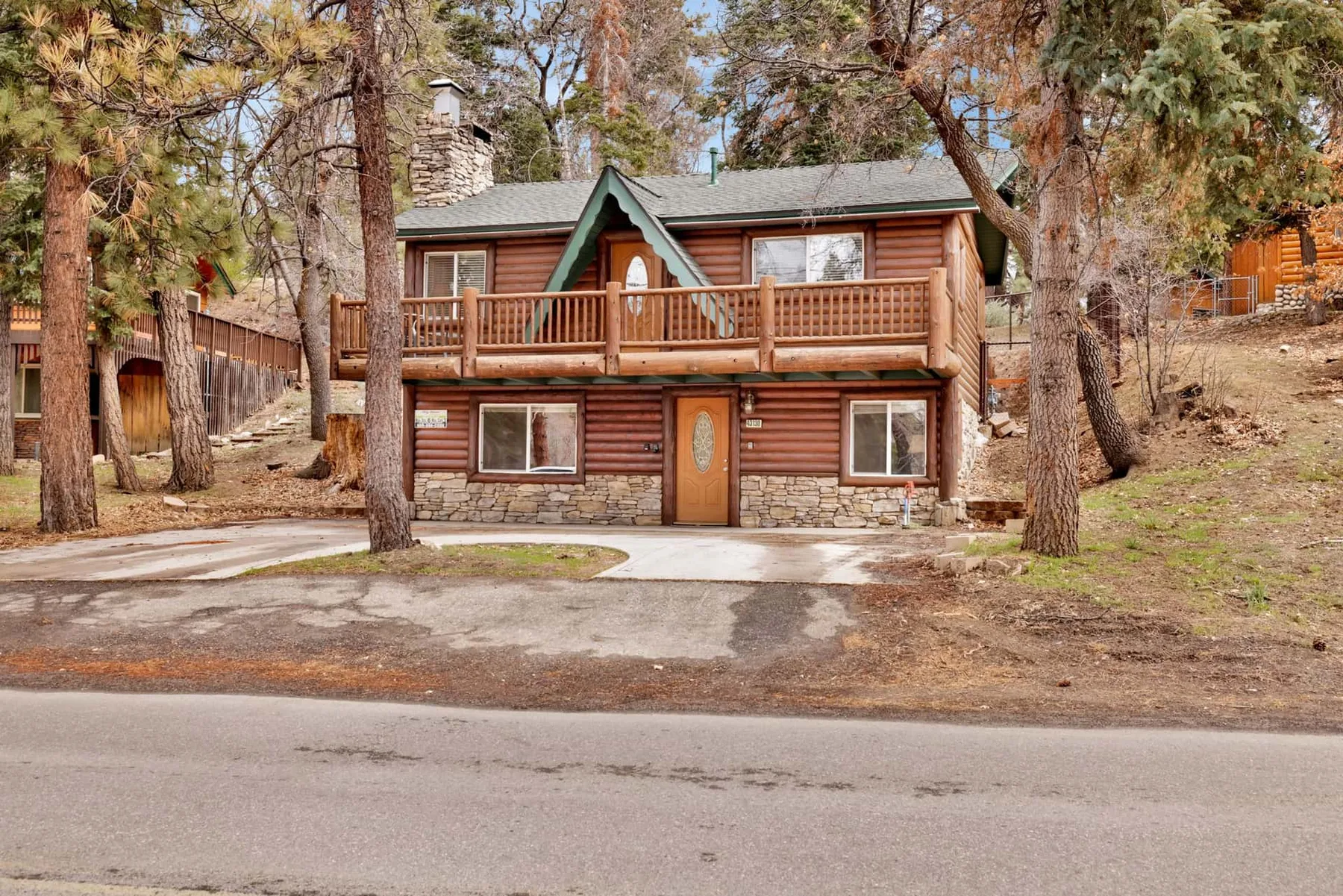 A two-story log cabin with a wooden balcony stands among trees beside a paved road.
