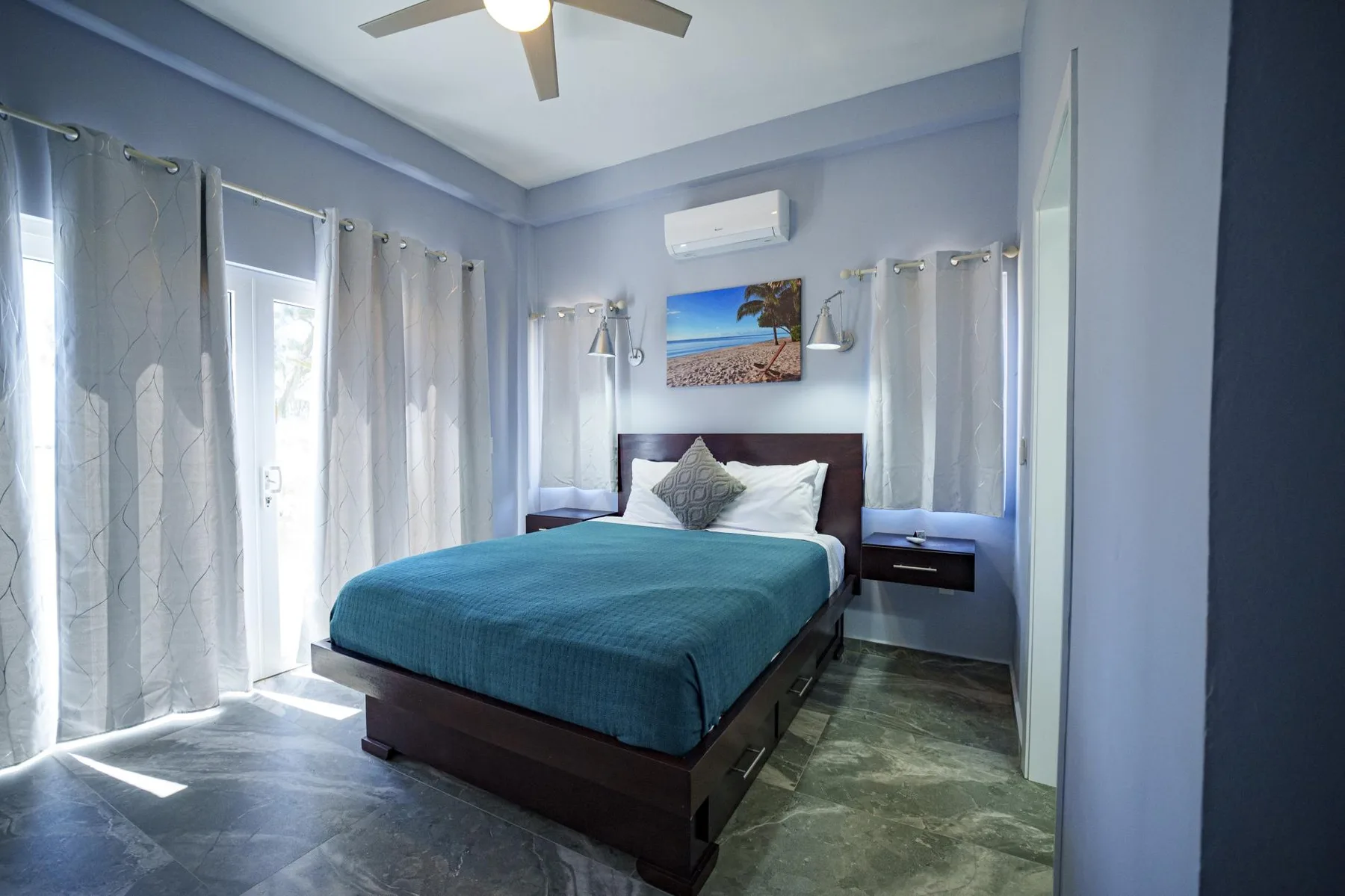A bedroom with a bed dressed in teal bedding and curtained windows.