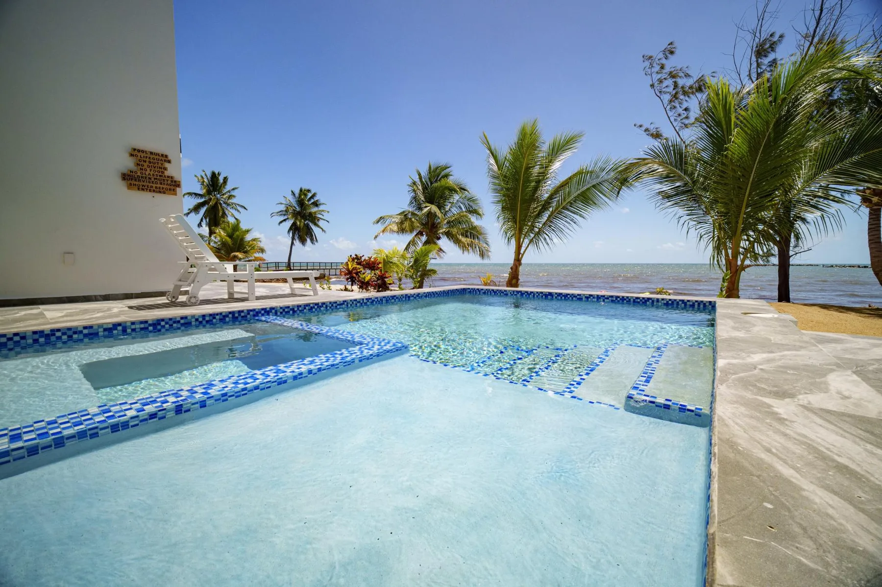 A shallow swimming pool beside a palm-lined beach and the sea.