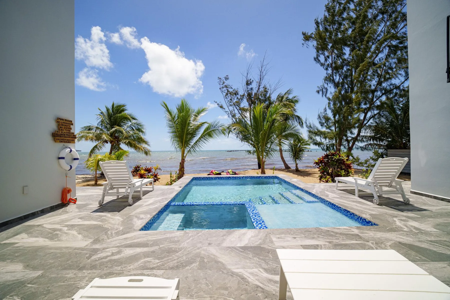 An outdoor swimming pool sits beside a sandy beach, with palm trees and lounge seating.