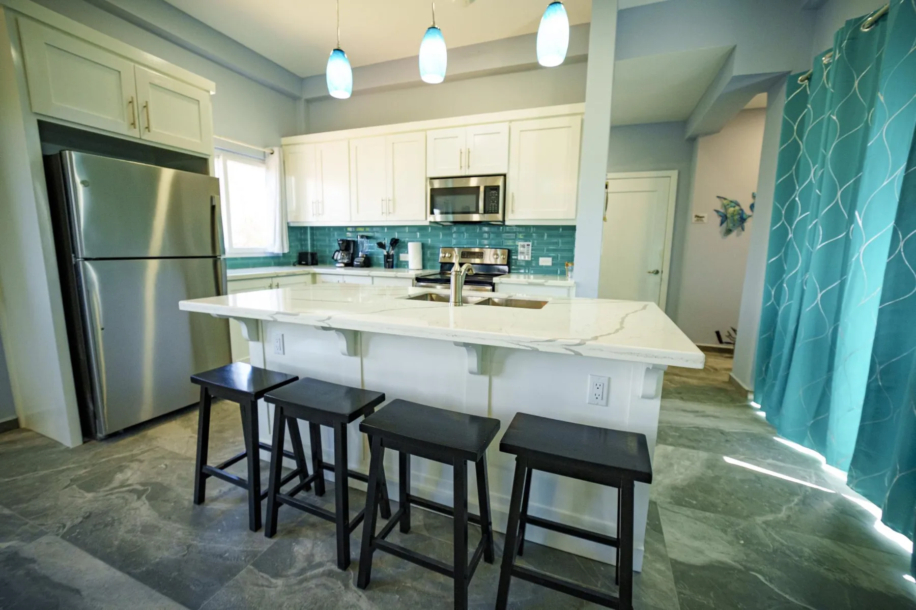 A bright kitchen with a large island, bar seating, and teal cabinetry.