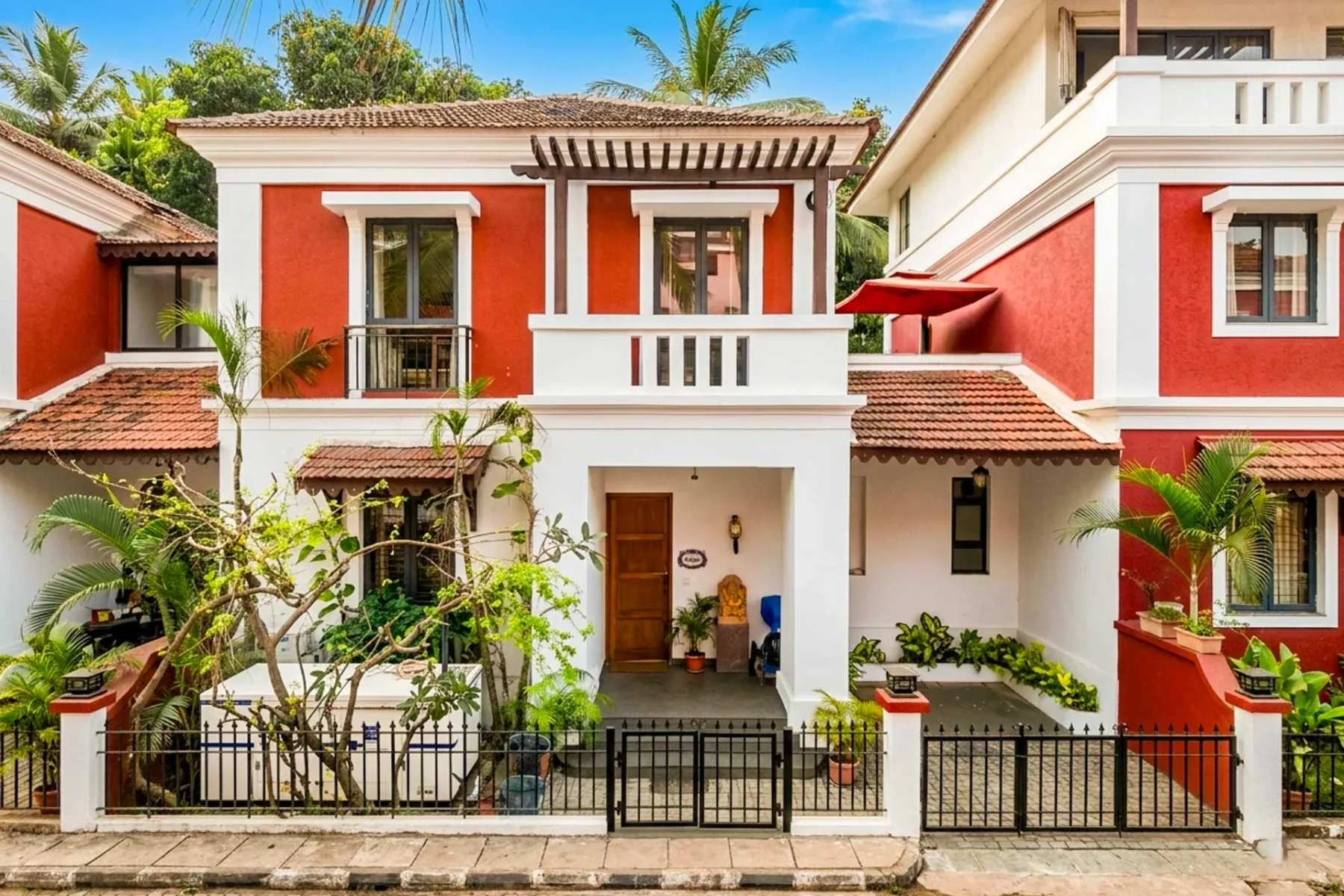 Un edificio rojo y blanco con entrada central, balcón y jardín delantero.