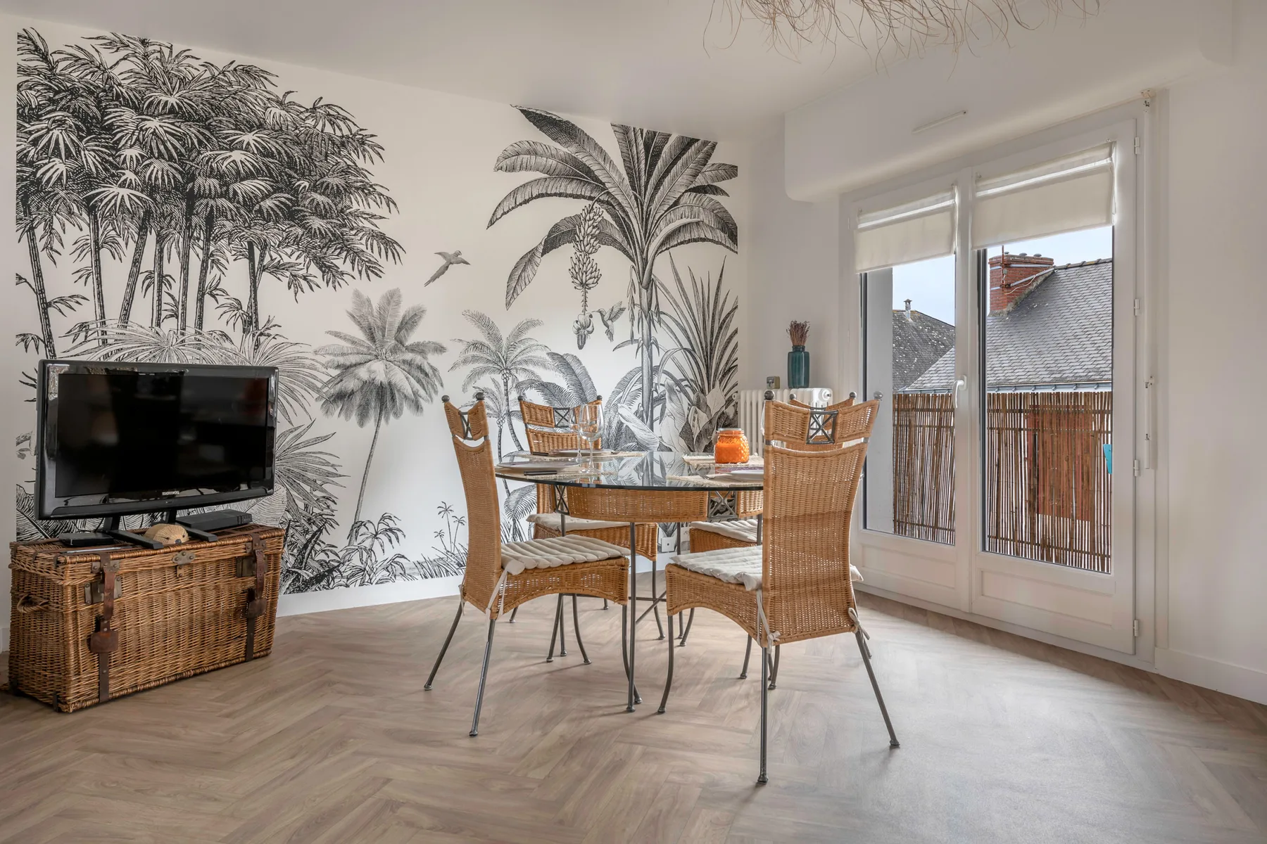 Un comedor con asientos de mimbre y un mural tropical junto a puertas de vidrio.