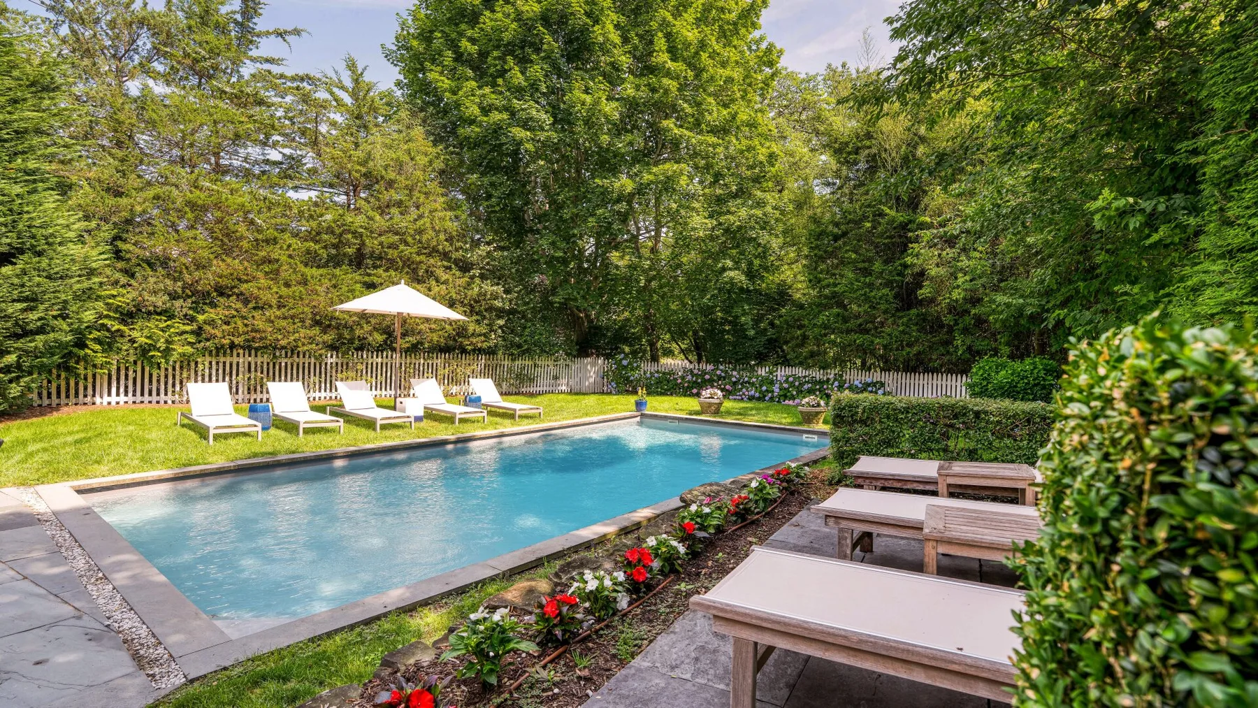 An outdoor swimming pool is bordered by lounge seating, flowers, and leafy trees.