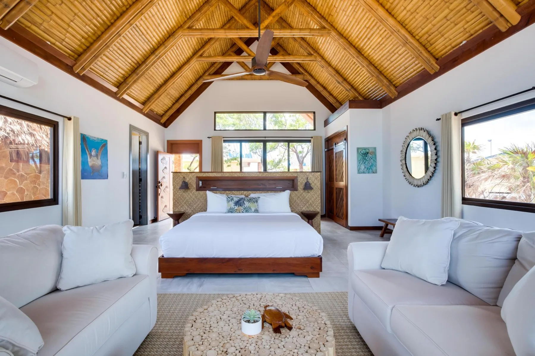 A bright bedroom with a bed between two seating areas beneath a high wooden ceiling.