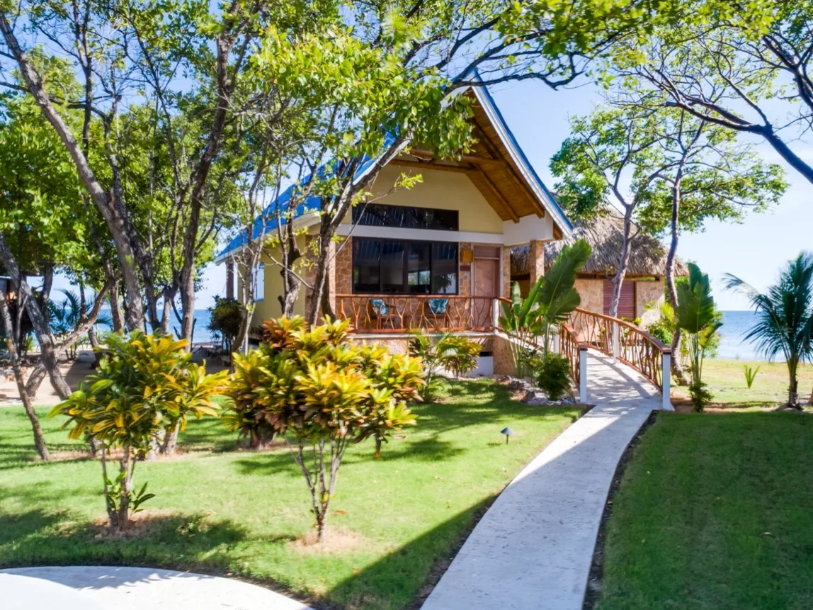 A small house with a peaked roof sits among trees beside a paved walkway.