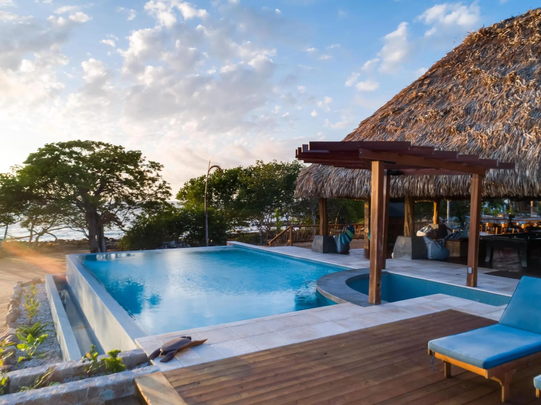 An outdoor pool beside a thatched-roof pavilion, with trees and sky beyond.