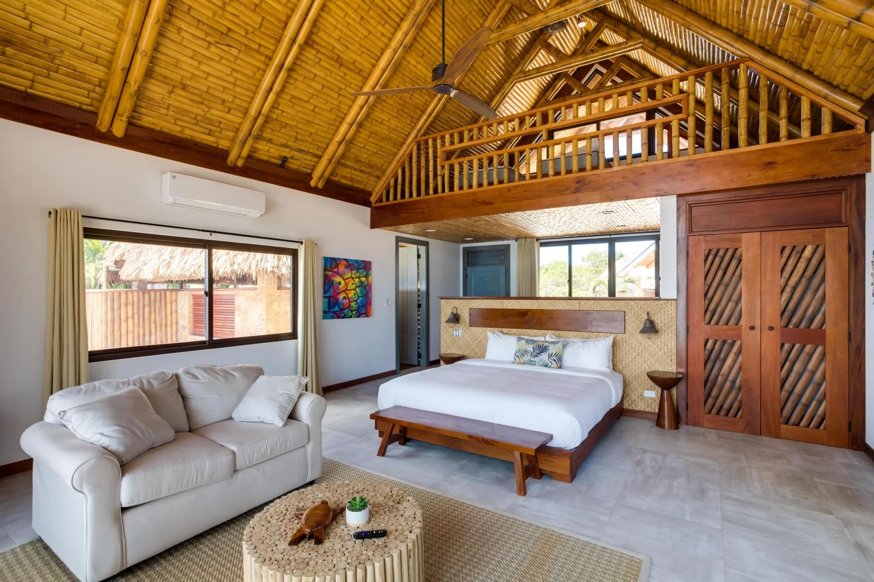 A bedroom with a bed, seating, and a wooden loft railing beneath a high ceiling.