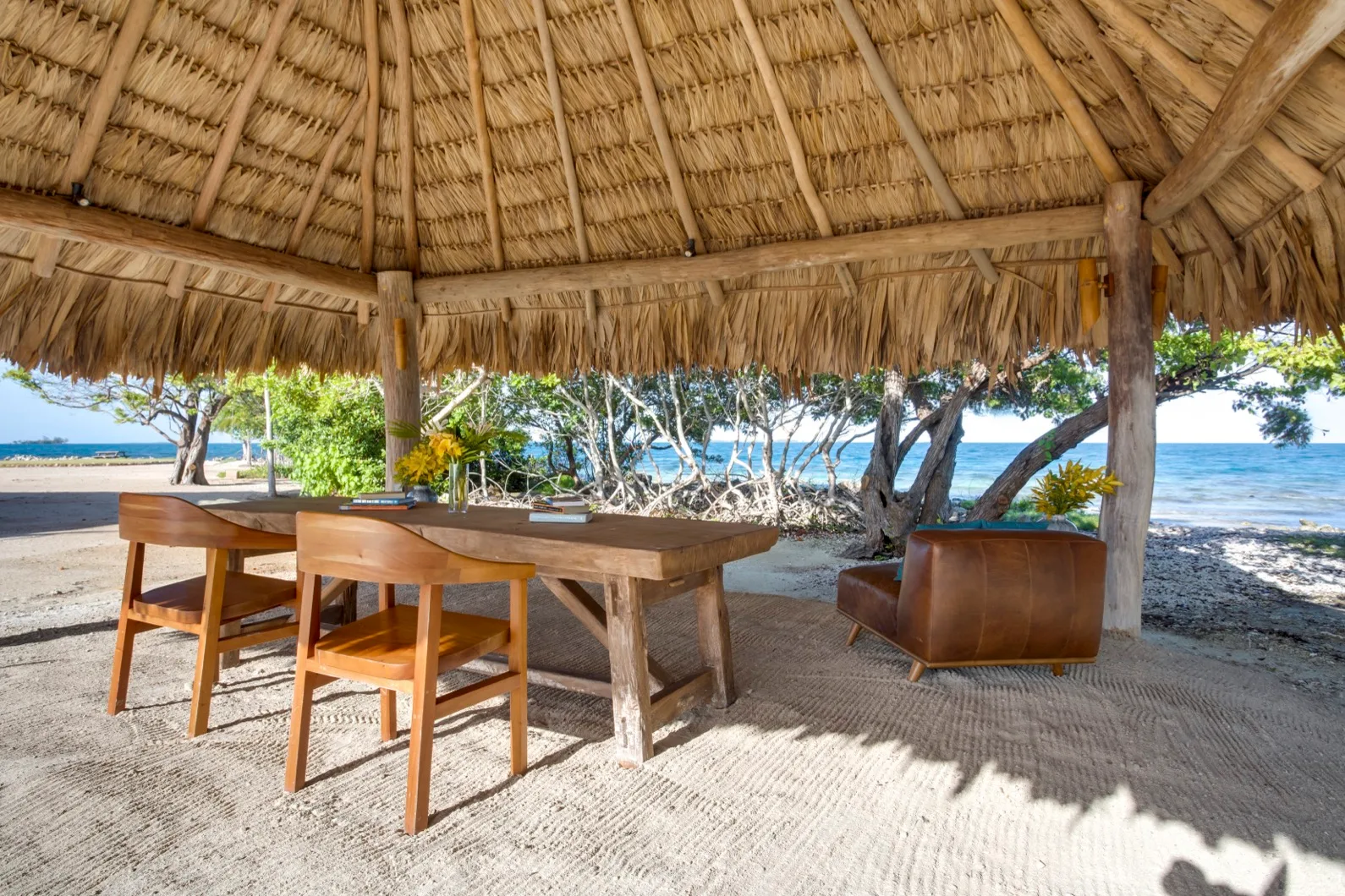 A thatched outdoor dining area with a wooden table and chairs beside the beach.